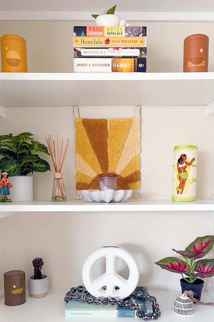 Shelf decorated with books, plants, candles, sculptures, hula girls, and tiki mug