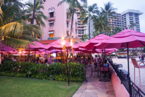 Outdoor tables covered by pink umbrellas, surrounded by lit tiki torches