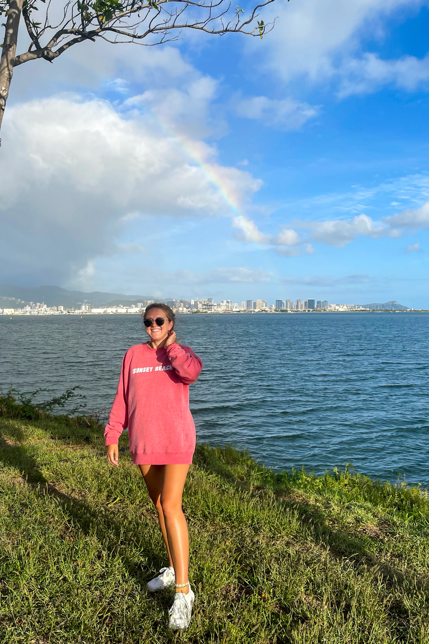 Lauren standing in front of rainbow over Waikiki in red sweatshirt, white sneakers and sunglasses